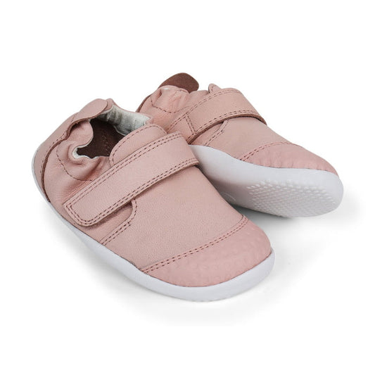 The Bobux Explorer Go Seashell Pink First Walker Crawler 501011B features pink baby shoes with white rubber soles and Velcro straps, designed for flexibility, shown side by side on a white background.