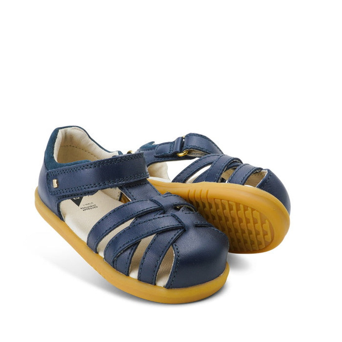 A pair of Bobux iwalk Cross Jump Sandal Navy Closed Toe 636703 kids' sandals with Velcro straps and tan rubber soles. One sandal stands upright, while the other is tilted to show the sole.