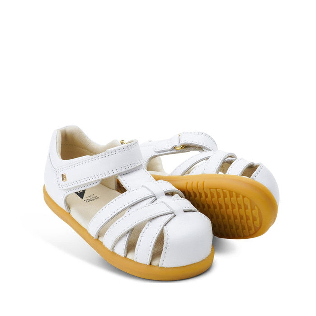 The Bobux iwalk Cross Jump Sandal White Closed Toe 636706 is a pair of children's white leather sandals with closed toes, adjustable Velcro straps, and light brown rubber soles, displayed on a white background.