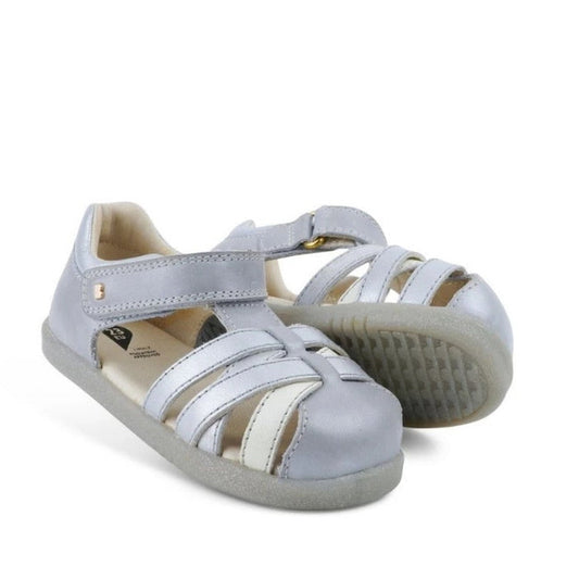 The Bobux iWalk Cross Jump Sandal Silver Pearl Closed Toe 636702 features kids’ leather sandals in silver and white with closed toes, velcro fastening, and rubber soles. One sandal stands upright, the other tilted to show the sole on a white background.