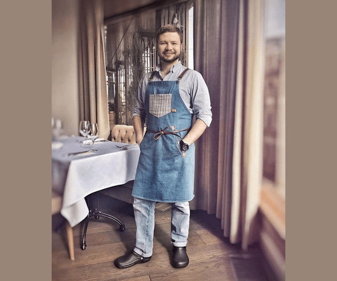 A smiling man in a blue apron, light shirt, jeans, and black Crocs Bistro Chefs Clog with Anti Slip Sole Unisex stands by a table set with glassware and a white tablecloth in a restaurant with large windows and curtains., 883503071094, 883503071100, 88350