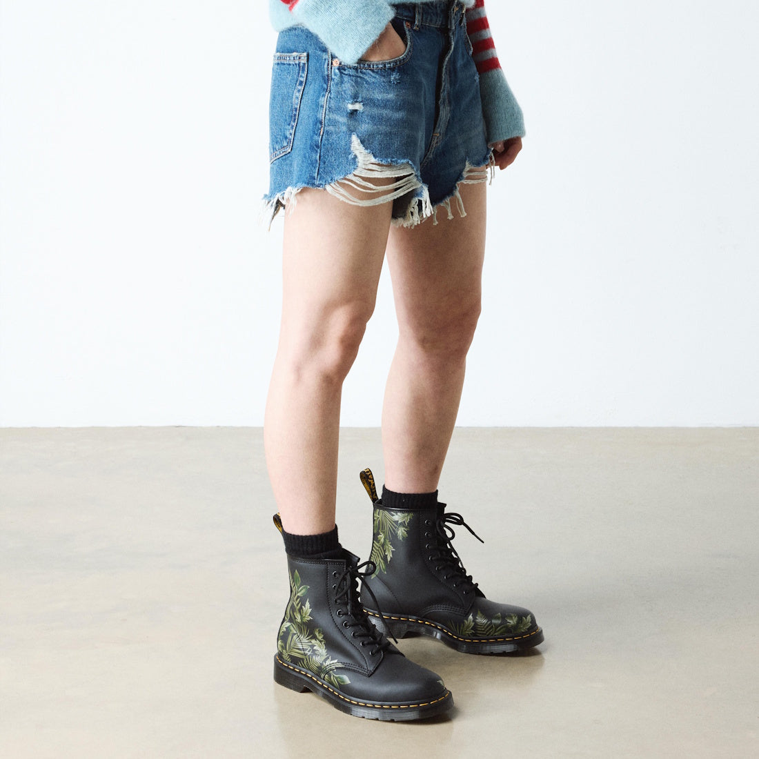 A person in a light blue sweater, frayed denim shorts, black socks, and 1460 Dr Martens Smooth EverGreen Leather 8 Up Boots with a green leaves pattern on a black background stands on a smooth, light-colored floor.