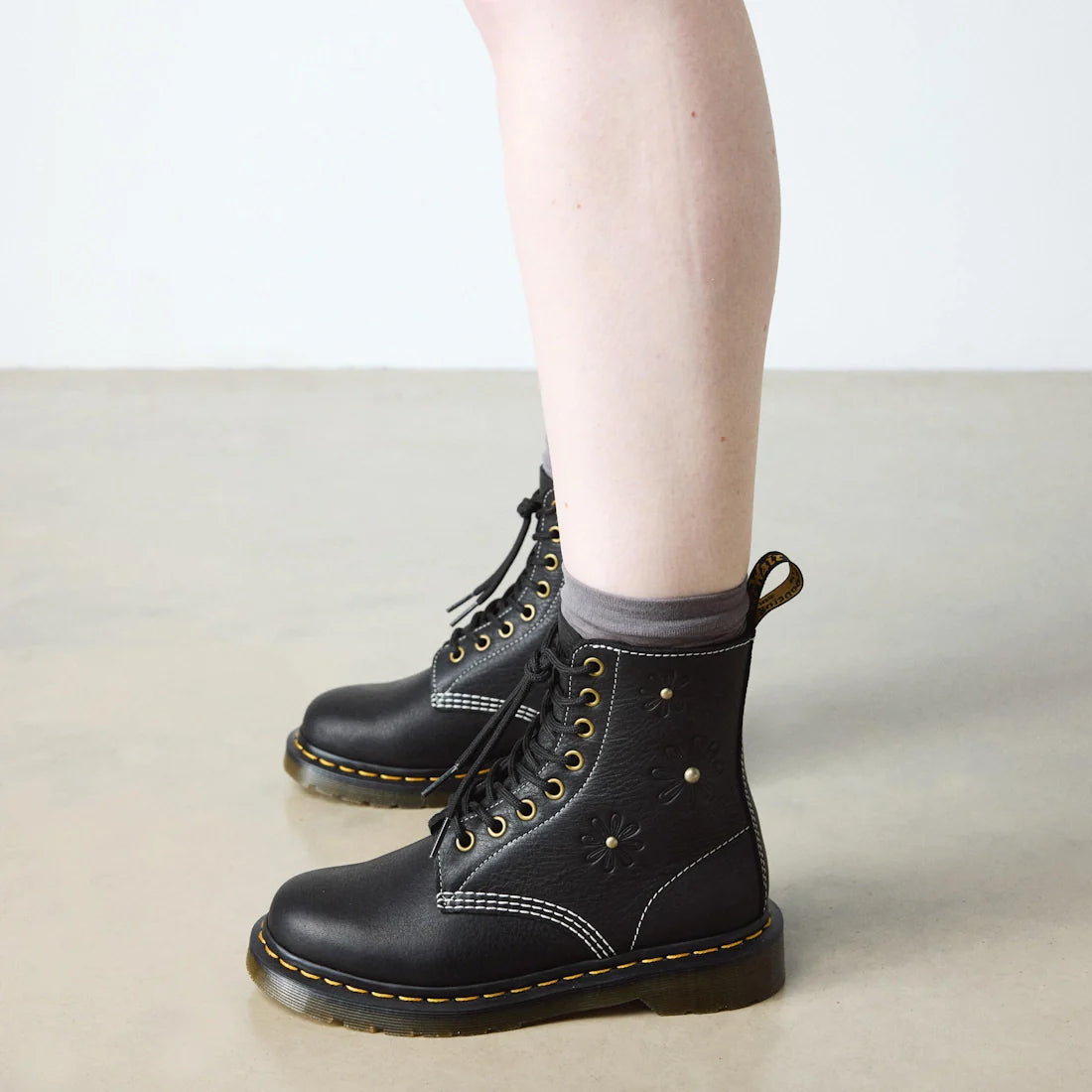 A person wearing black Dr. Martens Women's 1460 Pascal Flower Black Grizzly Leather Boots with floral details and gray socks stands on a light-colored floor against a plain white wall., 52216, 190665781137, 190665781144, 190665781151, 190665781175, 190665