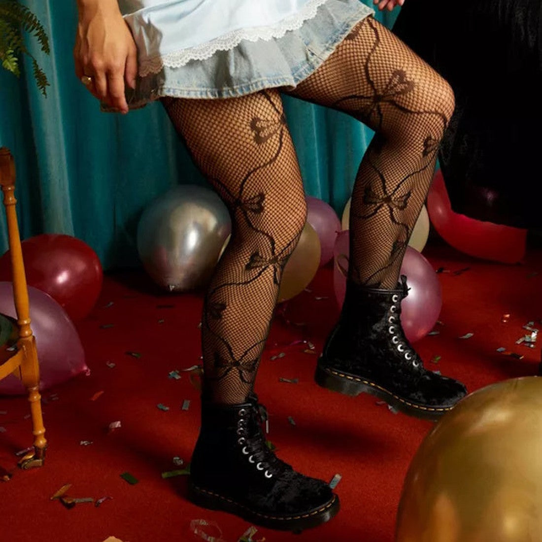 A person wearing a short denim skirt, black floral fishnet tights, and Dr Martens 1460 Vegan Black Crushed Velvet 8 Eye Boots stands on a red carpet with balloons, confetti, and a wooden chair., 190665580938, 190665580945, 190665580952, 190665580969, 1906