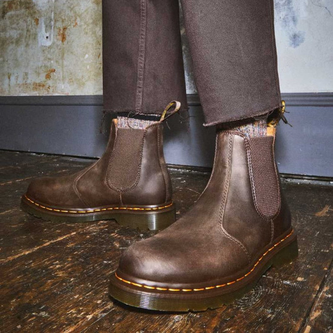 A person wearing Dr Martens 2976 YS Chelsea Boot Yellow Stitching Crazy Horse Brown with brown pants stands on a worn wooden floor near a scuffed wall and gray baseboard., 190665464962, 190665464979, 190665464986, 190665465006, 190665465013, 190665465020,