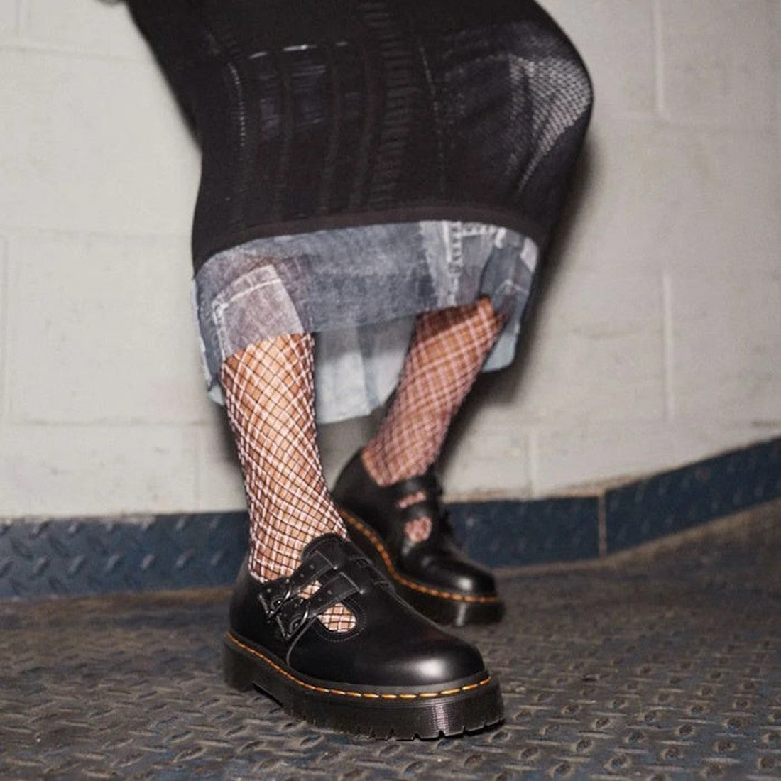 A person wearing a black skirt, fishnet stockings, and Dr Martens Bex 8065 II 2 Strap Mary Jane Shoes in black stands on a textured metal floor against a light-colored wall., 190665536799, 190665536805, 190665536812, 190665536829, 190665536843, 1906655368