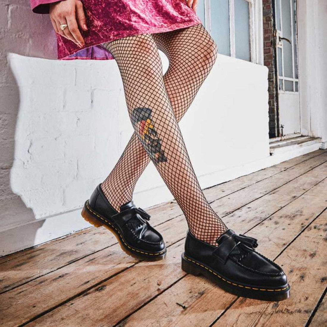 A person wearing fishnet tights with a colorful tattoo, a pink velvet skirt, and Dr Martens Adrian YS Black Smooth Tassel Loafer Shoes with Yellow Stitching stands on a wooden floor near a white wall and large windows., 22422, 883985989108, 883985989115, 