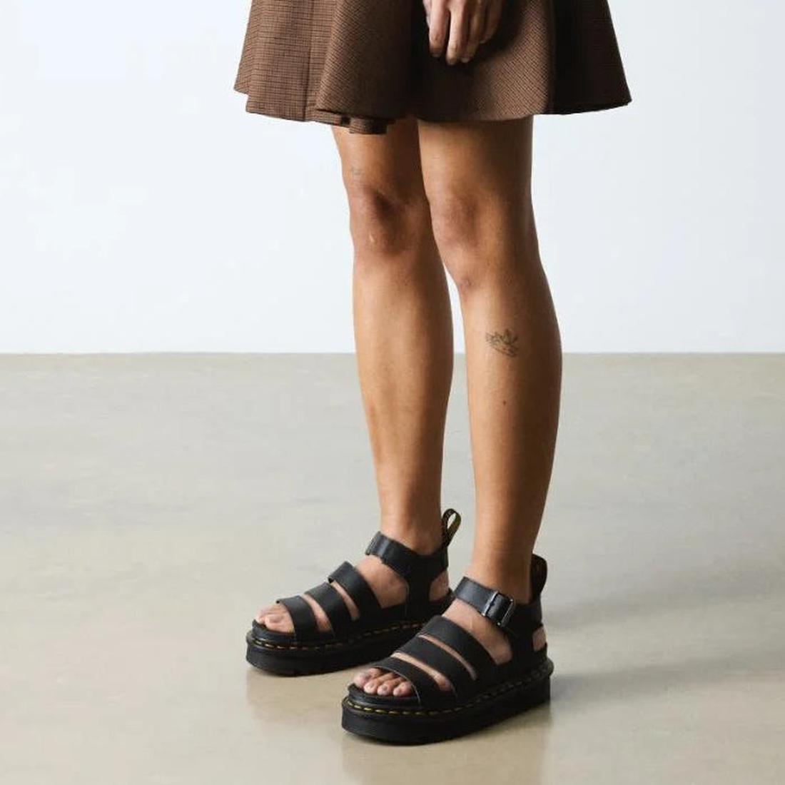 A womens standing side on to the camera wearing a brown mini skirt and Dr Martens Blaire 3 STrap Sandals in Athena Leather. Showing the strap and buckle around the ankle, yellow stitching and platform sole.,
190665725629,
190665725636,
190665725643,
19066