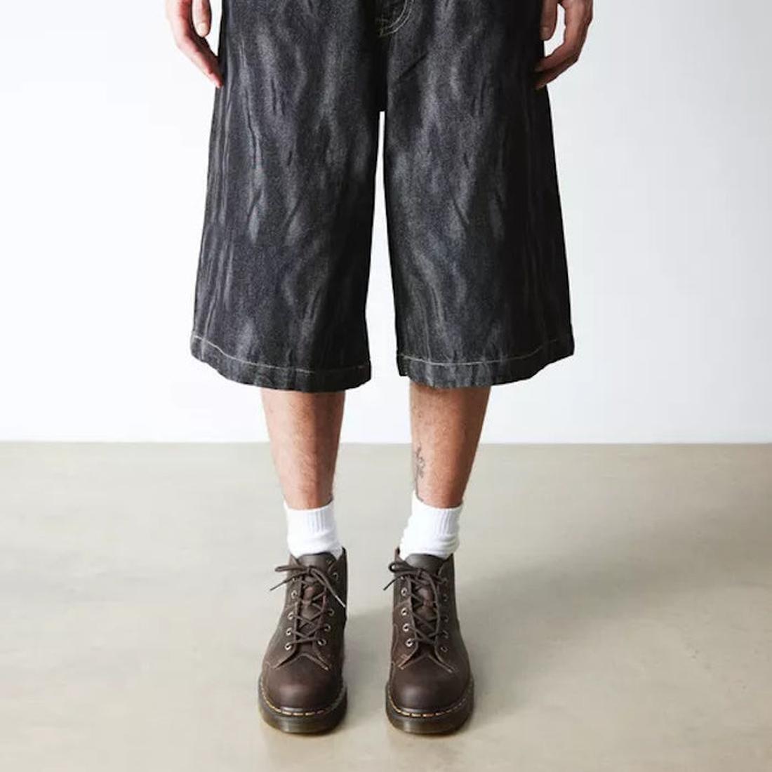 A person wearing long, loose black denim shorts, white socks, and Dr Martens Church Monkey Boot Crazy Horse Dark Brown stands on a light floor against a plain white wall. Only their legs are visible, highlighting a vintage-inspired look., 44507, 190665703