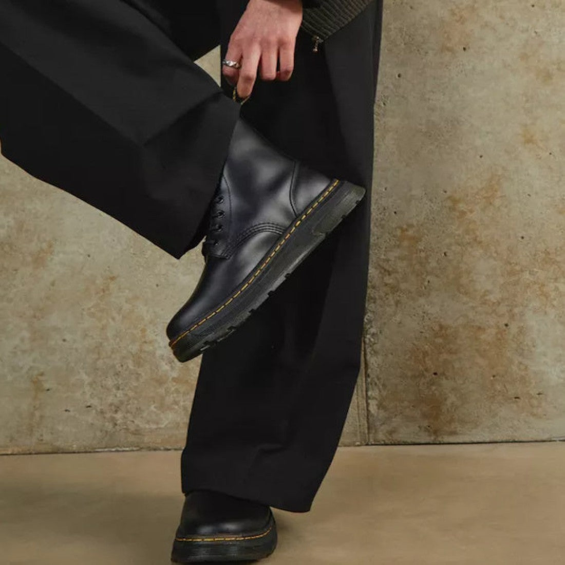 A person in loose black pants and Dr Martens Crewson Classic Leather Everyday Boots Black stands against a textured beige wall, lifting one leg to rest the boot on the opposite knee., 190665568400, 32121, 190665568417, 190665568424, 190665568448, 19066556