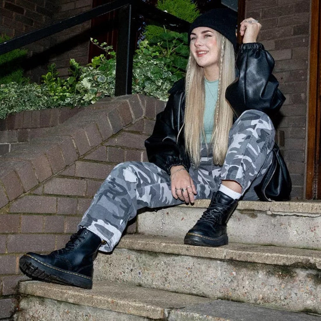 A woman with long blonde hair sits on outdoor steps, smiling with one hand raised. She wears a black beanie, black leather jacket, green top, gray camo pants, and Dr Martens Crewson Classic Leather Everyday Boots Black. Shrubs are in the background., 1906