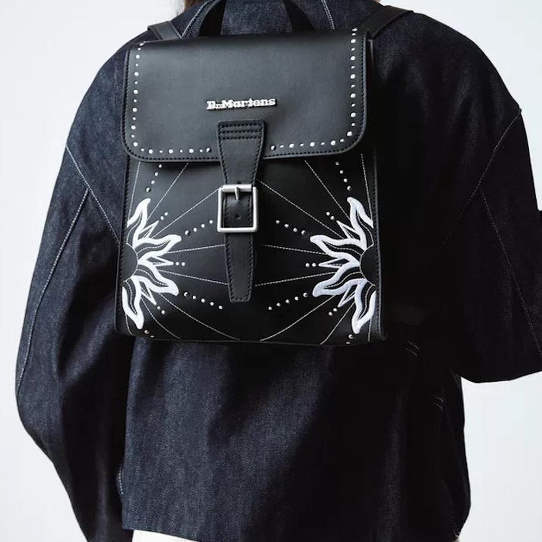 A person wearing a Dr Martens Mini Genix Nappa Solar Flare Leather Backpack Black with white sunburst designs and silver studs over a dark denim jacket. The bag features a front buckle closure., 190665739695