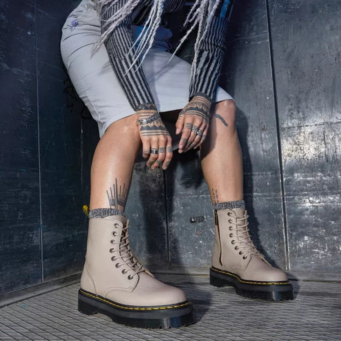 A person with tattooed hands and legs crouches in an industrial metallic setting, wearing a striped long-sleeve shirt, a light-colored skirt, and Dr Martens Jadon III Pisa Vintage Taupe Leather Boots., 190665590135, 190665590142, 190665590159, 32729, 1906