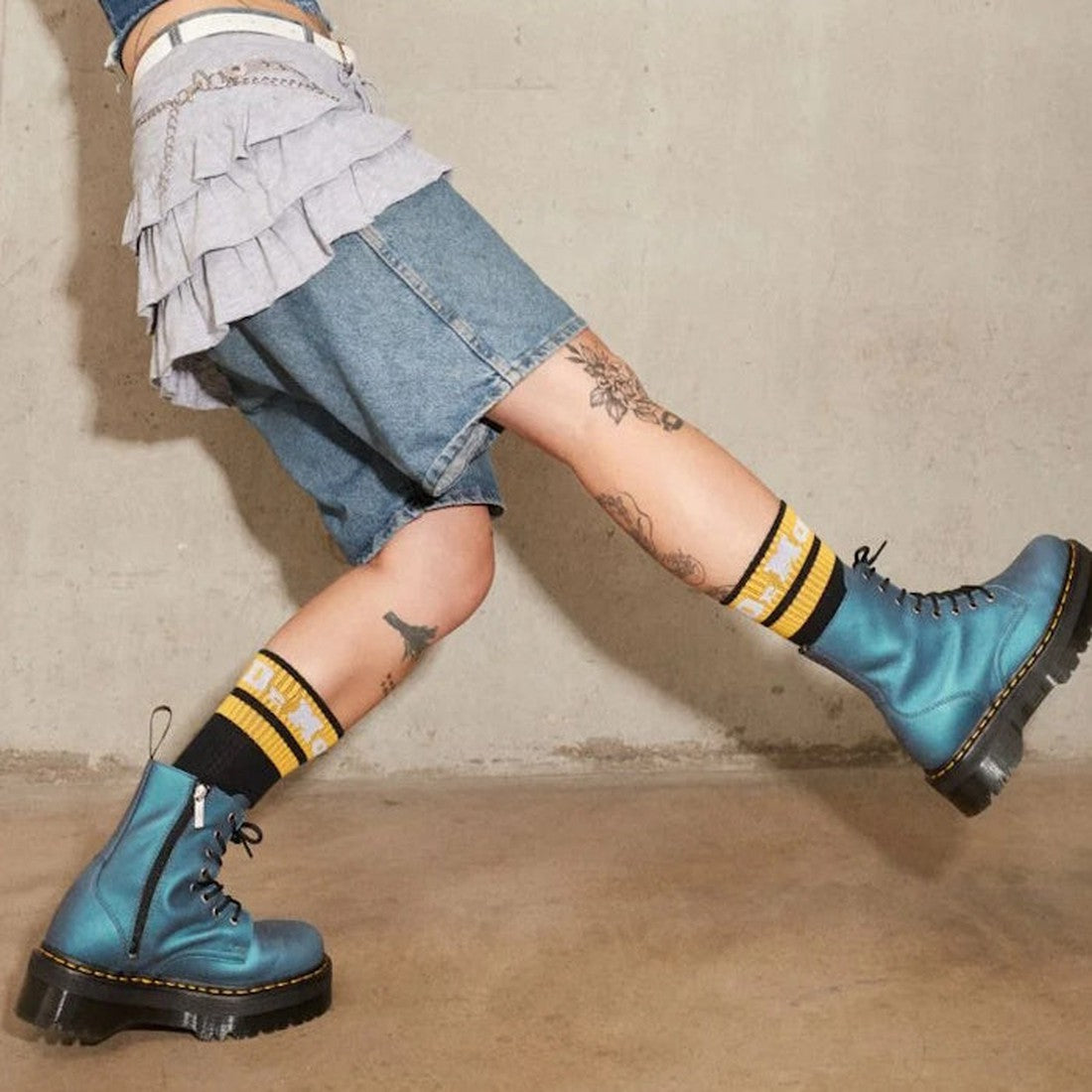 Person wearing blue denim shorts, a layered ruffle skirt, yellow-and-black socks, and Dr Martens Jadon Deep Blue Pull Up Leather Platform Boots with chunky tread, posing with one leg forward against plain concrete. Tattoos visible on both legs., 34144, 19