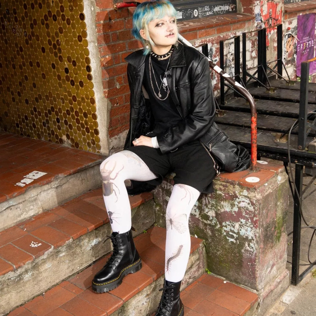 A person with short blue hair sits on outdoor steps, wearing a black leather jacket, black shorts, white tights with insect designs, layered necklaces, and Dr Martens - Jadon Polished Smooth Boot 8 up; graffiti and yellow tiles are in the background., 883