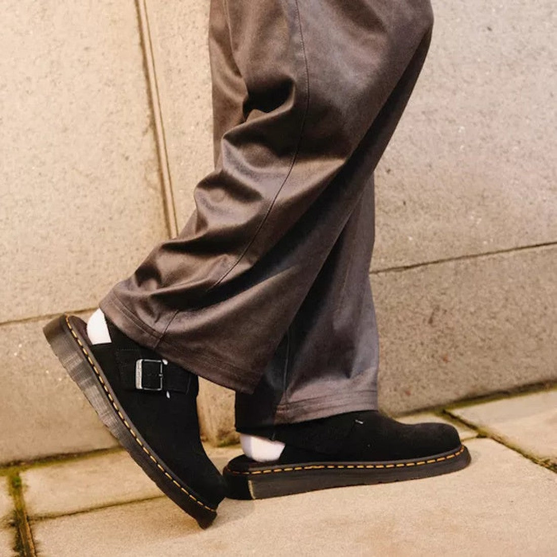 A person wearing loose gray pants and Dr Martens Black Jorge II Black Suede Sling Back Sandal Mules walks on a pavement next to a concrete wall., 190665645477, 190665645484, 190665645491, 190665645514, 190665645521, 190665645538, 190665645552, 19066564556