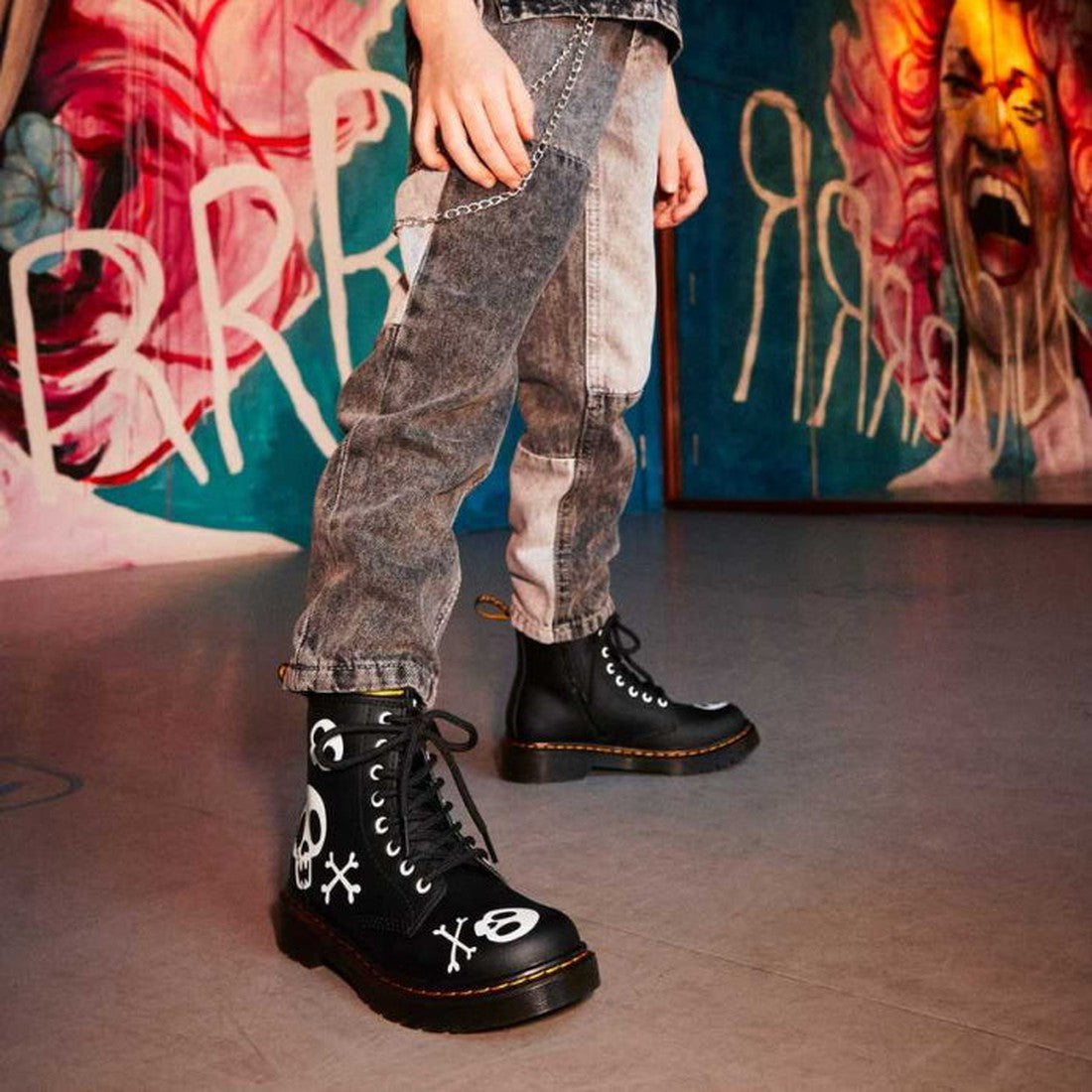 A kid in Dr Martens Kids 1460 Junior X Bones with Zip (UK 10), gray acid-wash jeans, and a chain stands before a vibrant mural of expressive faces and bold colors., 190665457018, 190665457032, 190665457056, 190665457063, 190665456967, 190665456981, 190665
