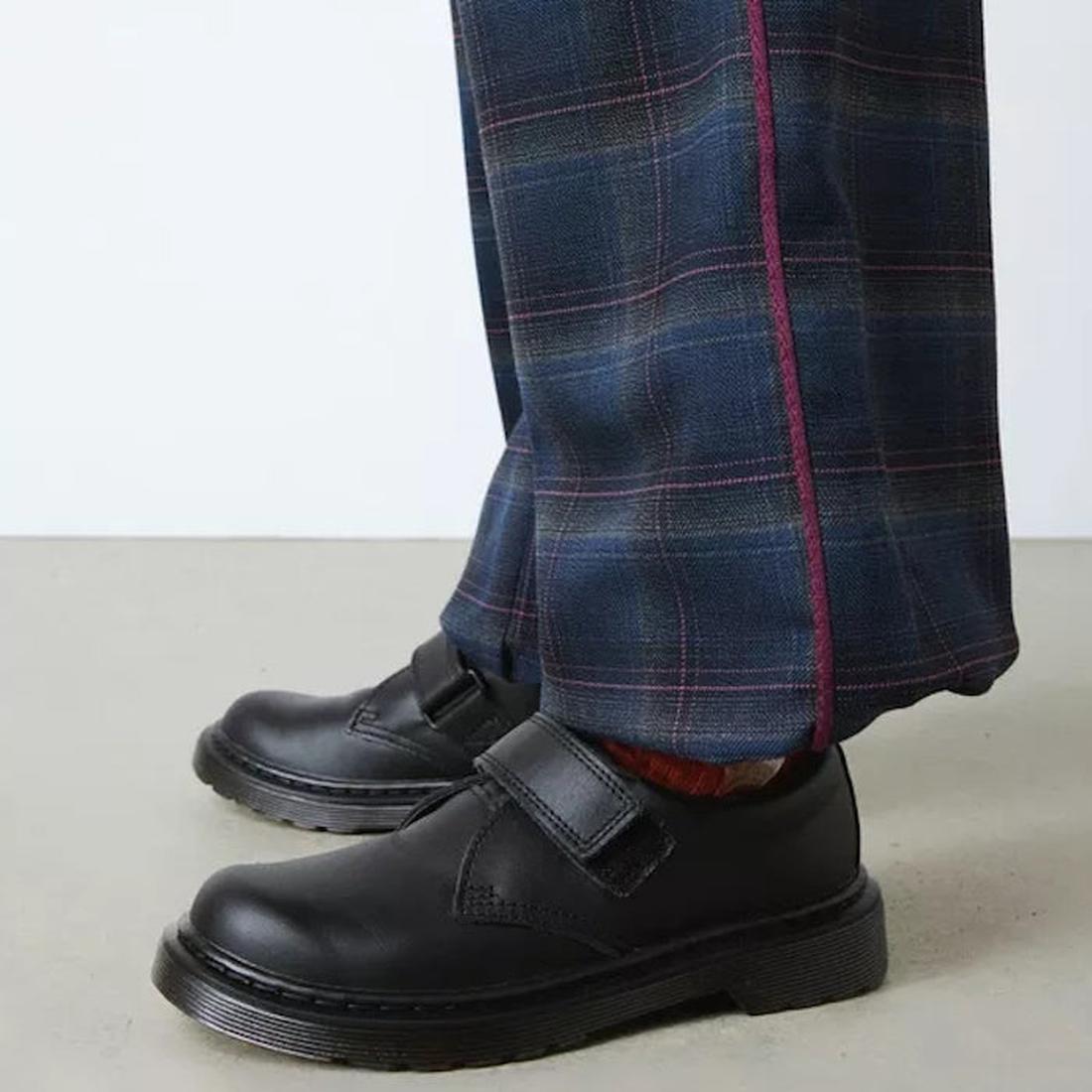A close-up of a person wearing blue plaid trousers with a maroon stripe, paired with Dr Martens Kids Kamron II Mono Velcro Junior Black School Shoes (UK 10-3), standing on a light gray floor., 190665681185, 190665681208, 190665681222, 190665681239, 190665