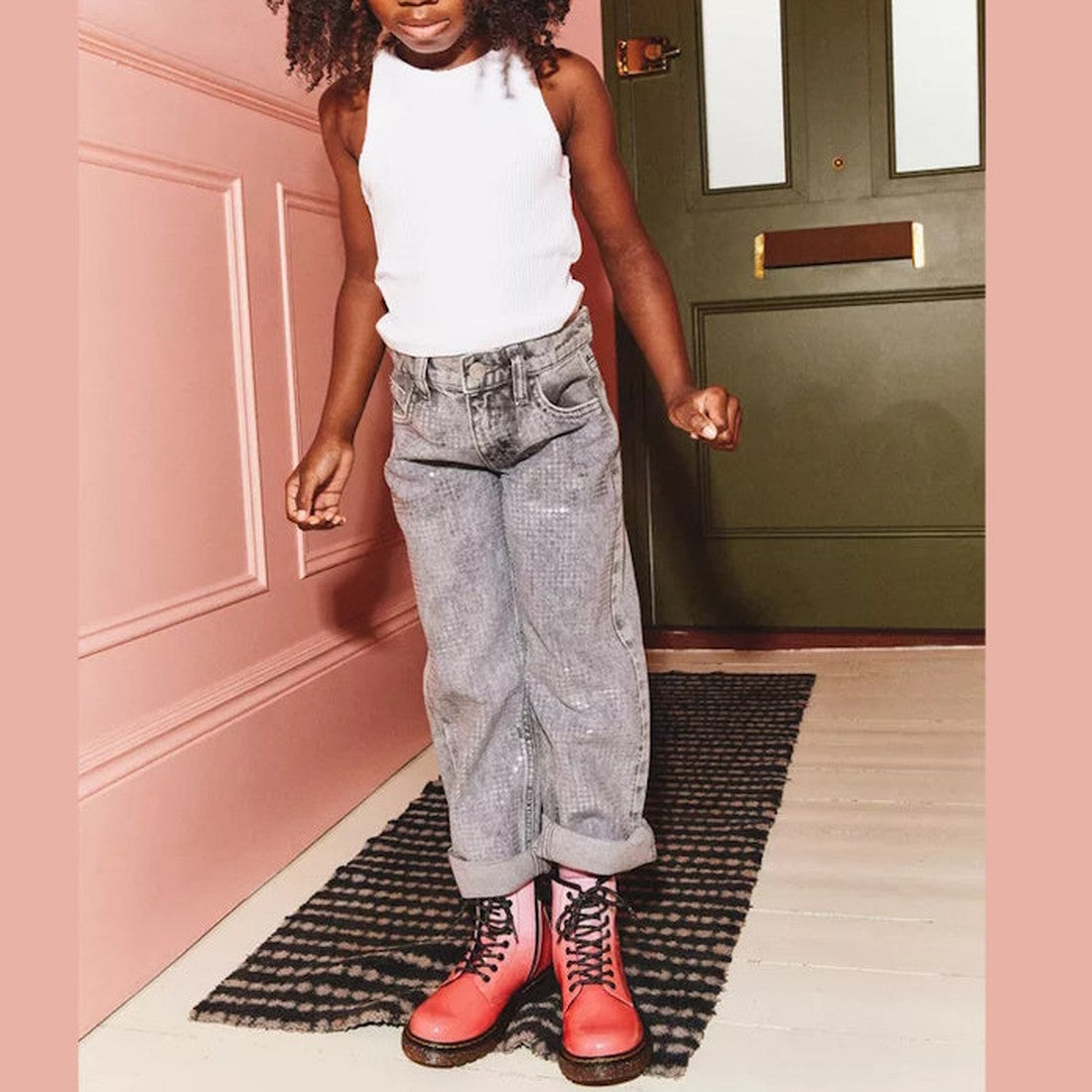 A child stands on a patterned rug by a green door, wearing a white tank top, gray cuffed jeans, and Dr Martens Kids 1460 Junior Pink Glitter Ombre Gradient boots, with one arm slightly bent and their face partially out of frame., 190665615463, 190665615364, 190665615388, 190665615401