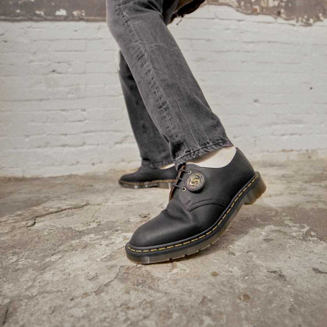Person wearing Dr Martens Made In England Oil Leather 1461 3 Eye Shoes with yellow stitching and a round gold emblem on the laces, paired with grey jeans, standing on a worn concrete floor by a white brick wall., 190665425826, 190665425895, 22989, 22986, 
