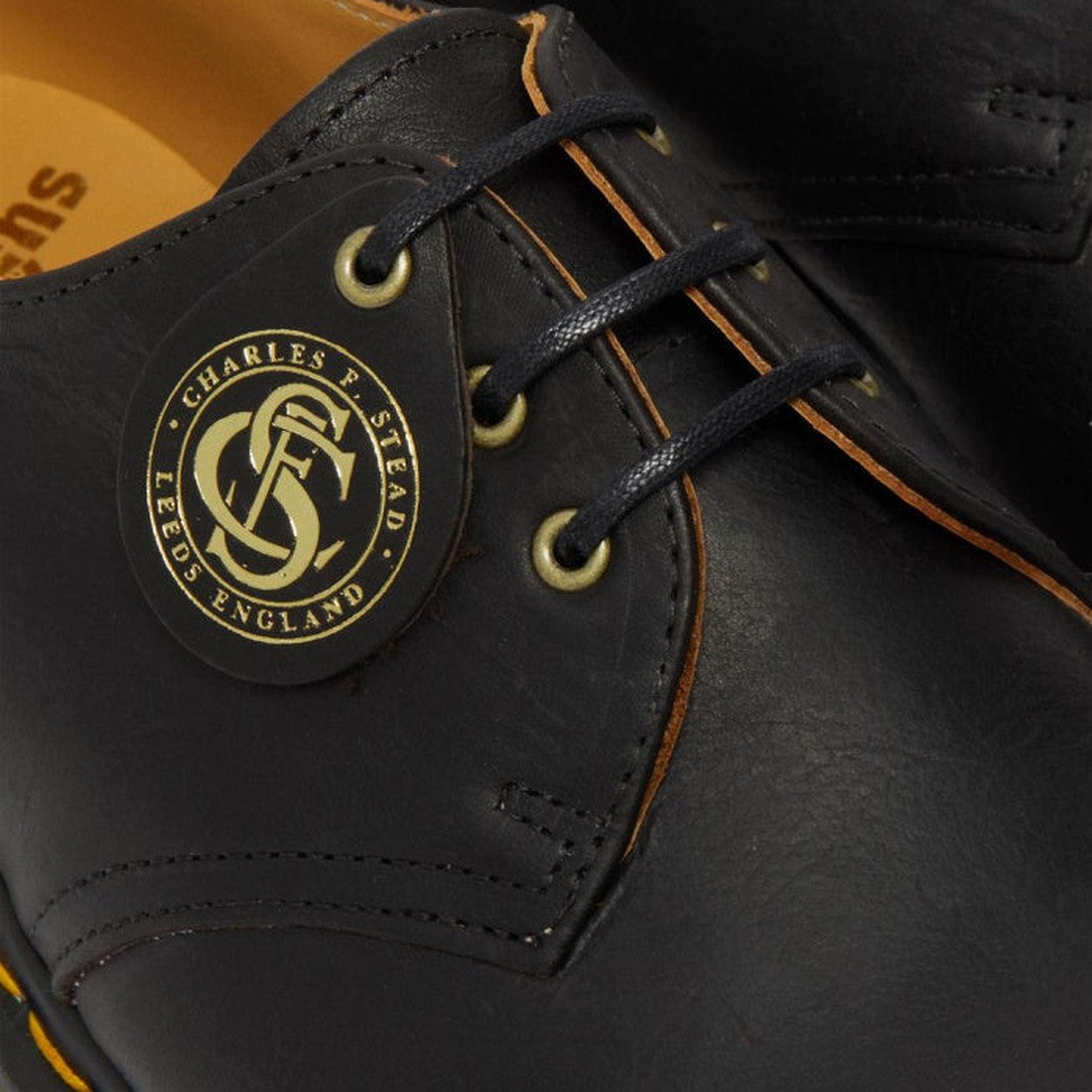 Close-up of Dr Martens Made In England Oil Leather 1461 3 Eye Shoes in black with gold eyelets and black laces. A round tag reads Charles F. Stead Leeds England with a monogram, highlighting its English craftsmanship., 190665425826, 190665425895, 22989, 2