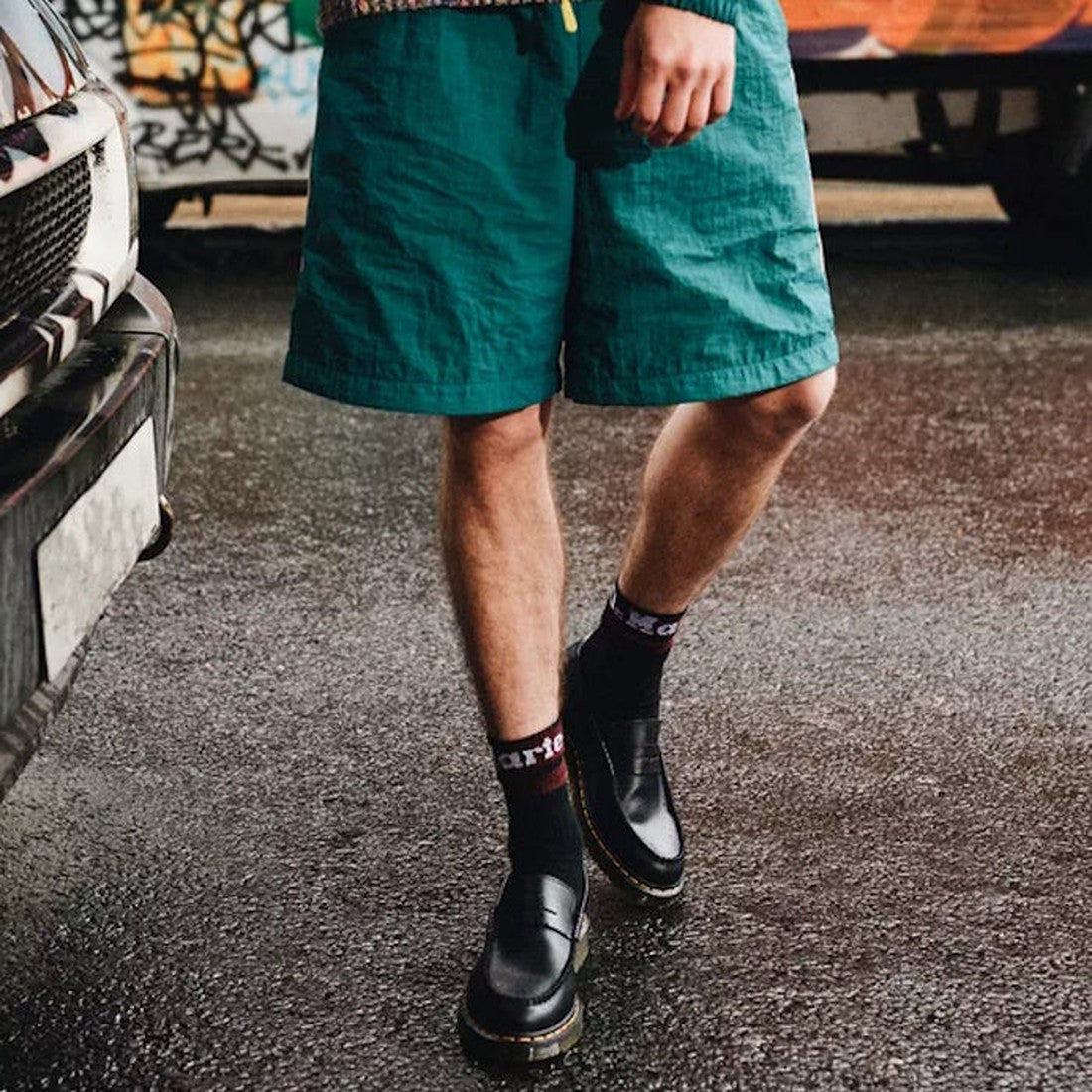 A person in green shorts, black socks with white and red lettering, and Dr. Martens Loafer Penton Slip On Black Smooth Leather Mens stands on a wet street near a white car and a graffiti-covered wall., 190665570434, 190665570502, 190665570519, 19066557044