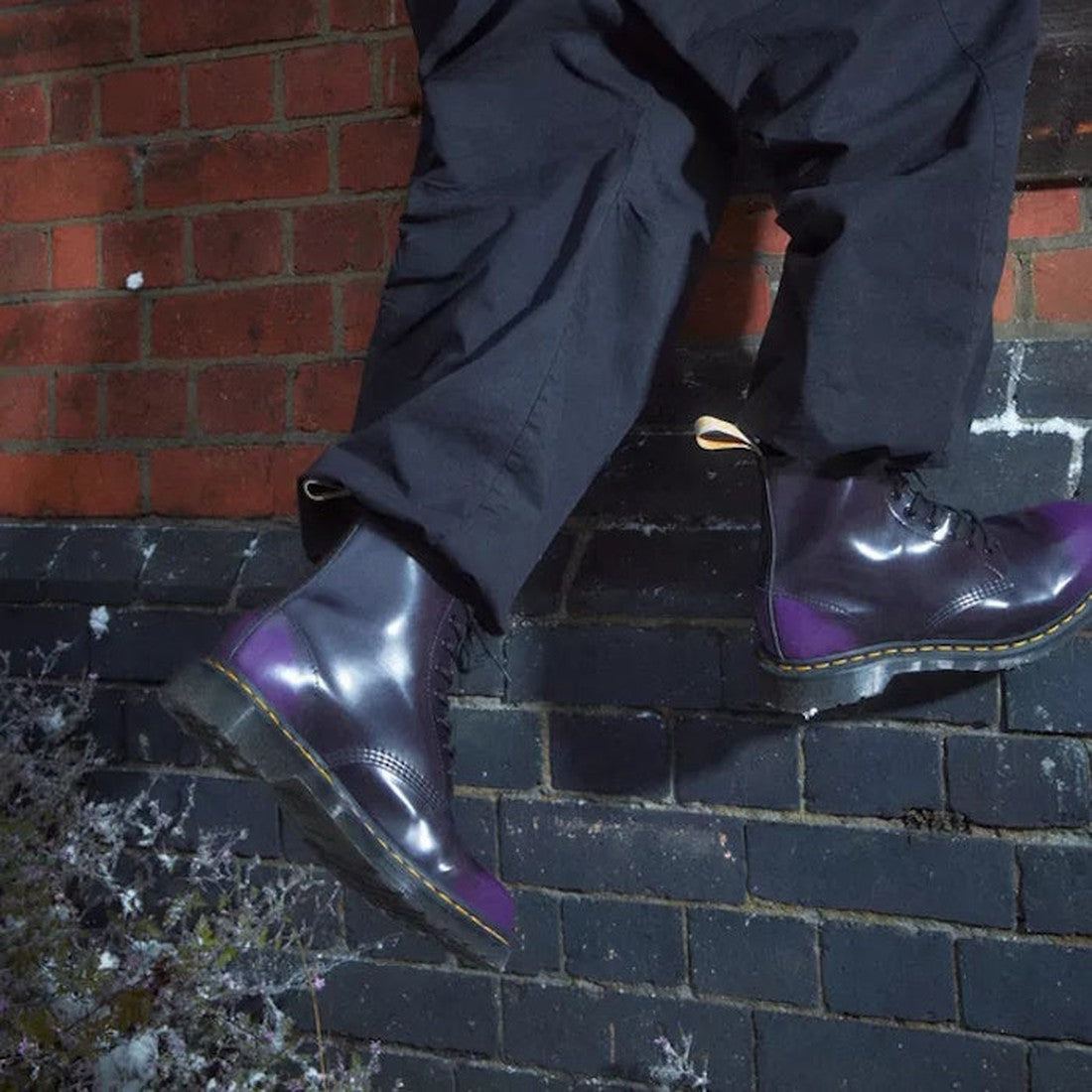 Wearing black pants and Dr. Martens Vegan 1460 Rich Purple Oxford Rub Off Boots, someone steps onto a brick wall at night—one foot on the wall, the other on the ground., 190665602845, 31837, 31838, 31839, 190665602852, 190665602869, 190665602883, 19066560
