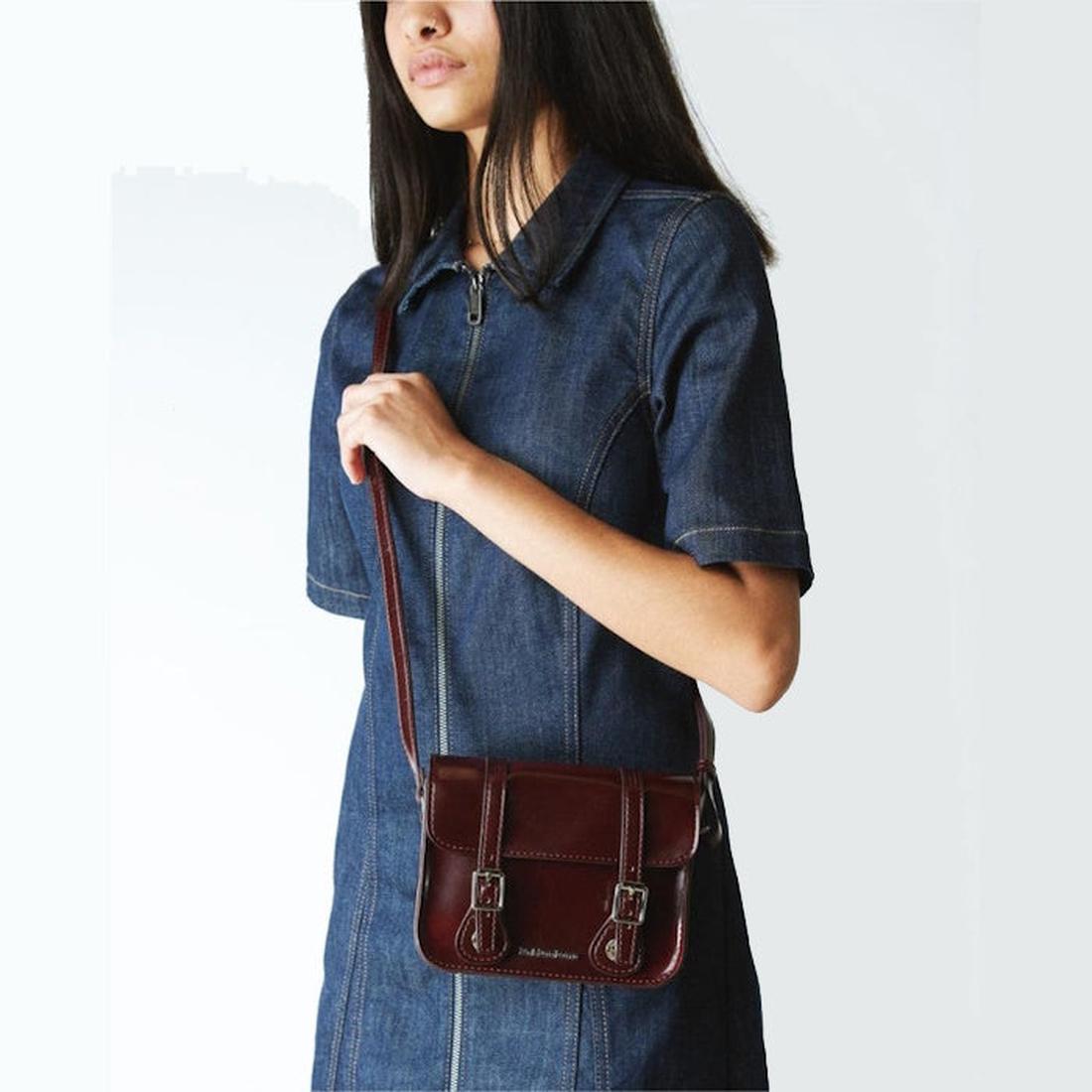 A woman in a short-sleeve denim dress holds the Dr Martens 7 Inch Satchel Vegan Cherry Red Oxford Brush, a durable everyday bag with buckle details, against a plain white background., 190665192087