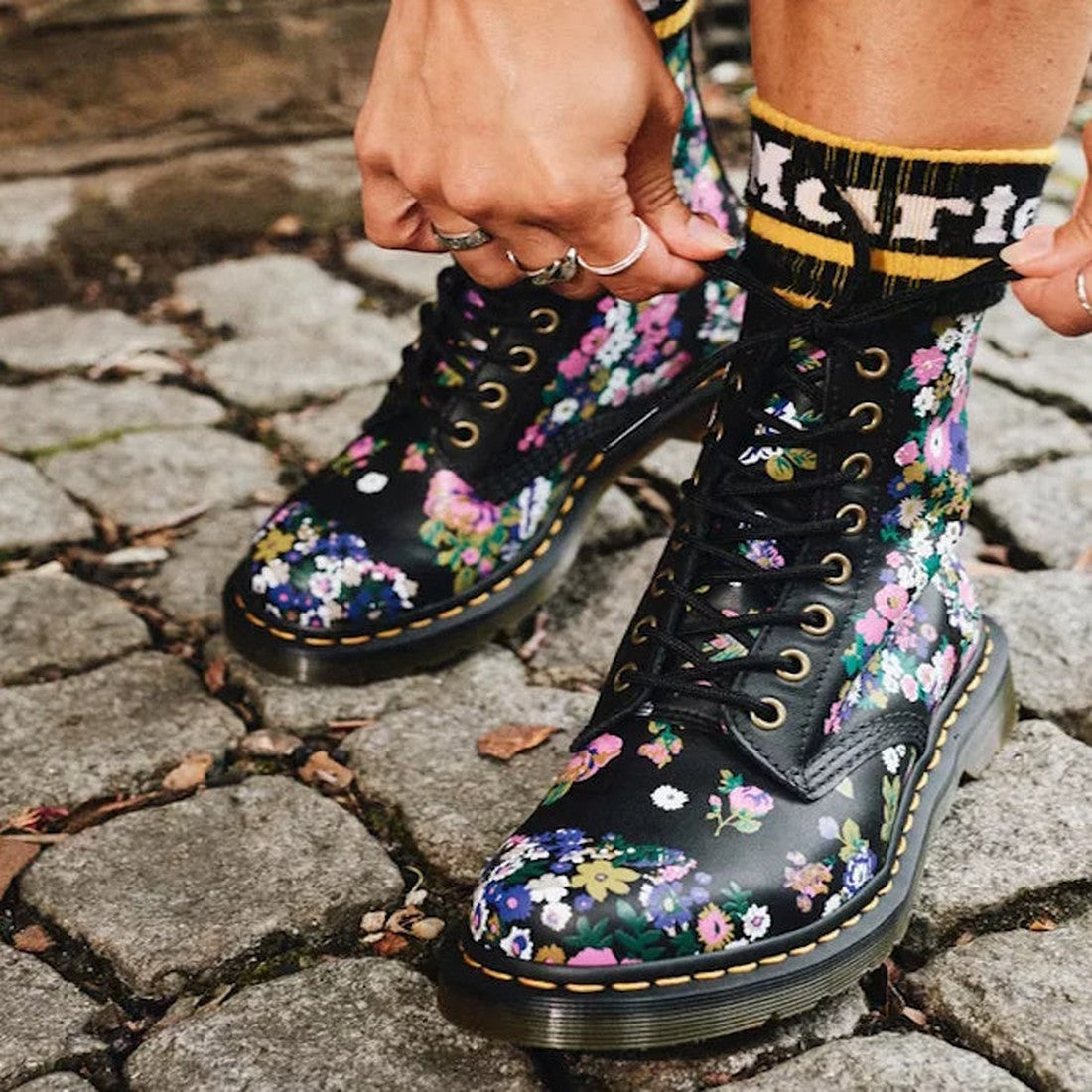 Someone laces up Dr Martens Vintage Floral 1460 Pascal Backhand Leather Multi boots on a cobblestone path, wearing black socks with yellow and white writing., 190665572490, 190665572506, 190665572513, 190665572537, 190665572544, 190665572551