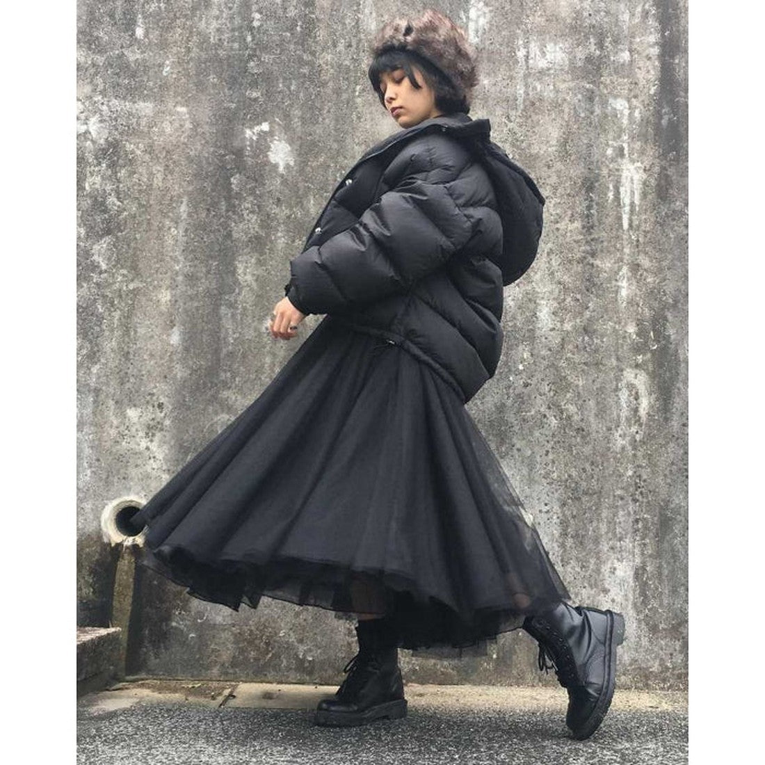 A person in a black puffer jacket, black tulle skirt, Dr Martens 1460 Black MONO 8 Up Boot Smooth, and a furry hat stands before a textured concrete wall, turning slightly as their skirt flows out., 883985396401, 883985396418, 883985396425, 883985396449, 