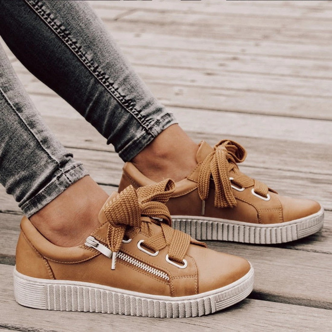 A person stands on a wooden deck in fitted gray jeans, wearing EOS Jovi Leather Trainer Tan sneakers with thick laces, white platform soles, and side zippers—casual leather shoes that blend style and elegance effortlessly., 10040, effegiejovitan137, effeg