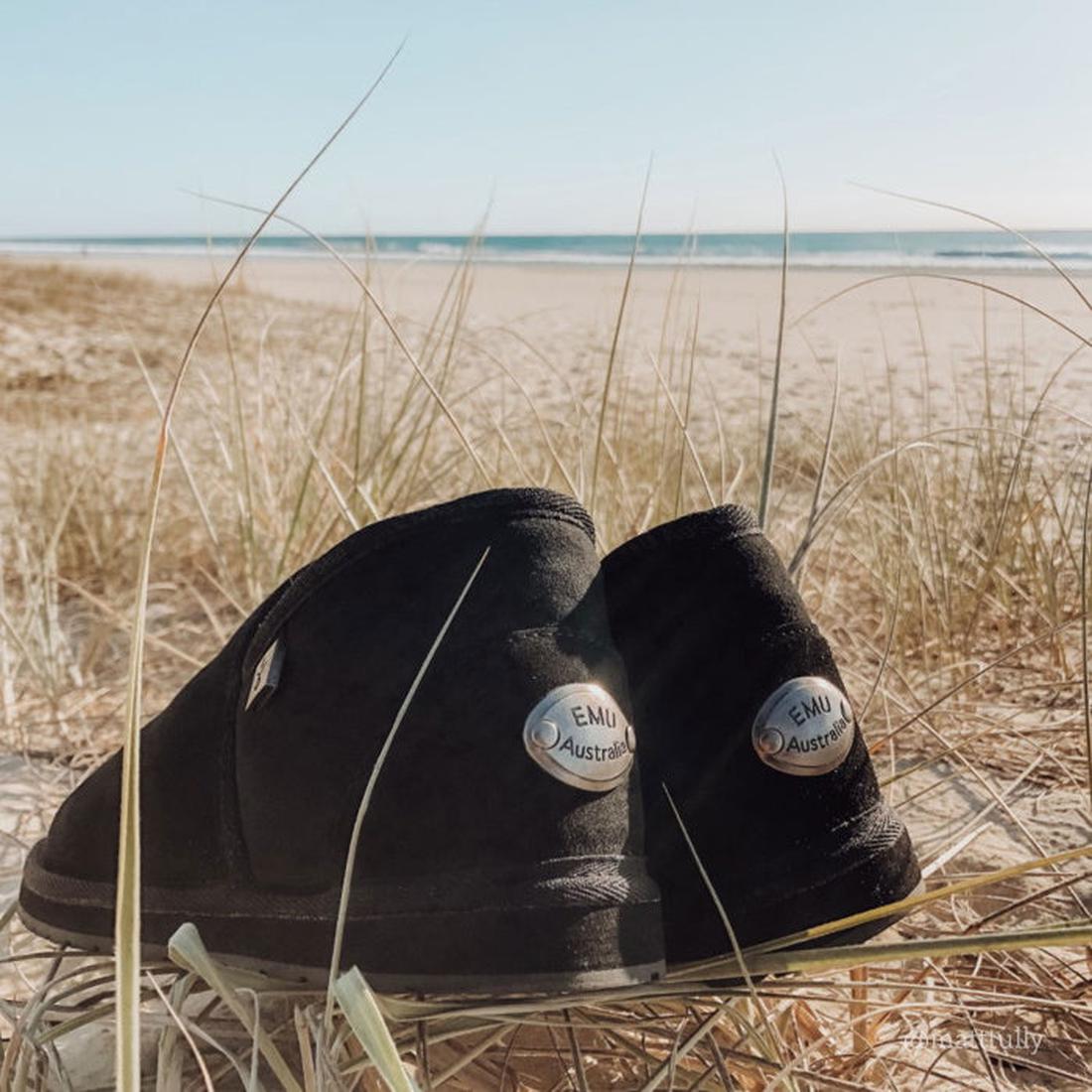 EMU NZ Ashford Slip On Sheepskin Unisex Slippers in black with cozy linings and sturdy soles sit in beach grass by sand and ocean.  ksnz