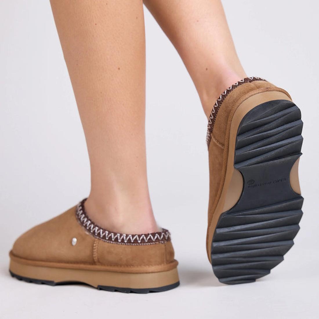 Tan EMU Sheepskin Outback Mule Sandl slippers on a modal looking from the back view in a walking poise showing the chunky serrated black sole  on a light gray background.,
9330071968499,
9330071968505,
9330071968512,
9330071968529,, 45249, 9330071968499, 