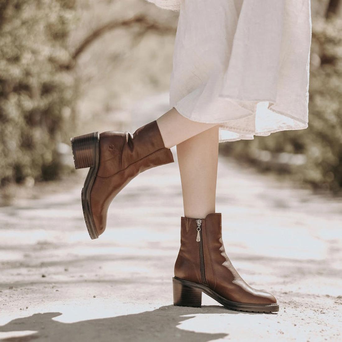 A model wearing a pair of EMU womens brown leather EMU Zeil ankle boots with a medium height block heel. The right foot is raised showing the outer side of the boot and the left foot is on the ground showing the inner zip., 9330071964217,
9330071964224,
9