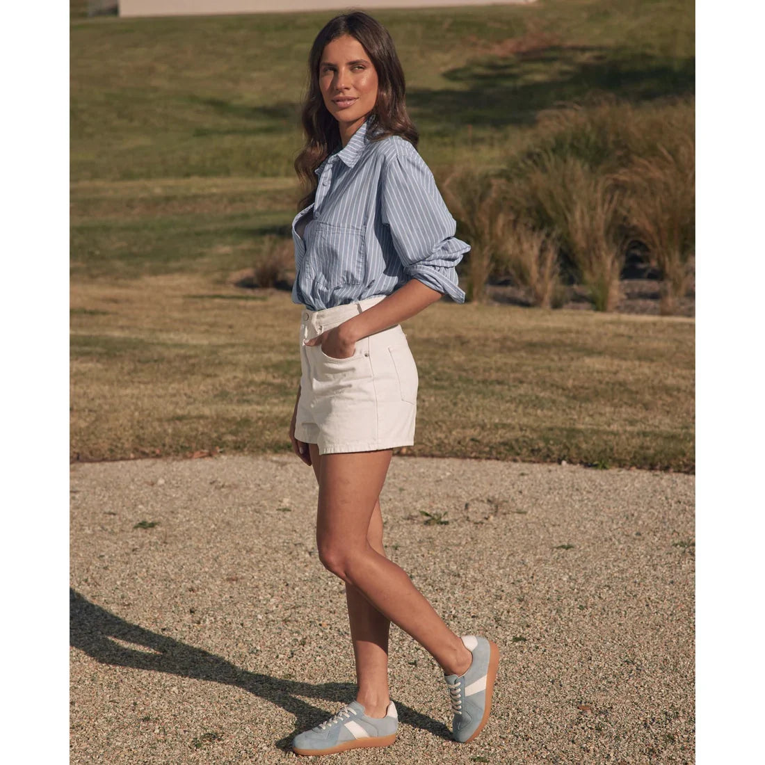 Outdoors on a gravel path, a woman in a blue striped shirt and white shorts faces the camera with one hand in her pocket, wearing EOS Melbaa Sky Suede Leather Lace Up Trainers; grassy lawn and shrubs are visible behind her., 41698, 9321191224558, 93211912