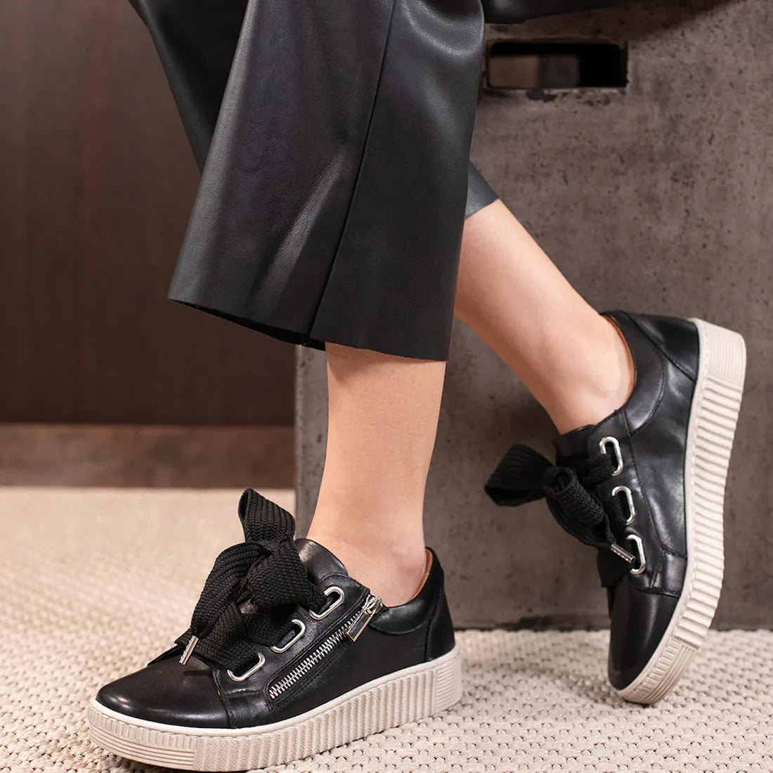 A person wearing black leather pants and EOS Joyce Leather Trainer Black Lace Up with Zip sneakers, featuring thick soles, large black laces, anatomical insoles, and side zippers, rests their feet on an indoor concrete surface.