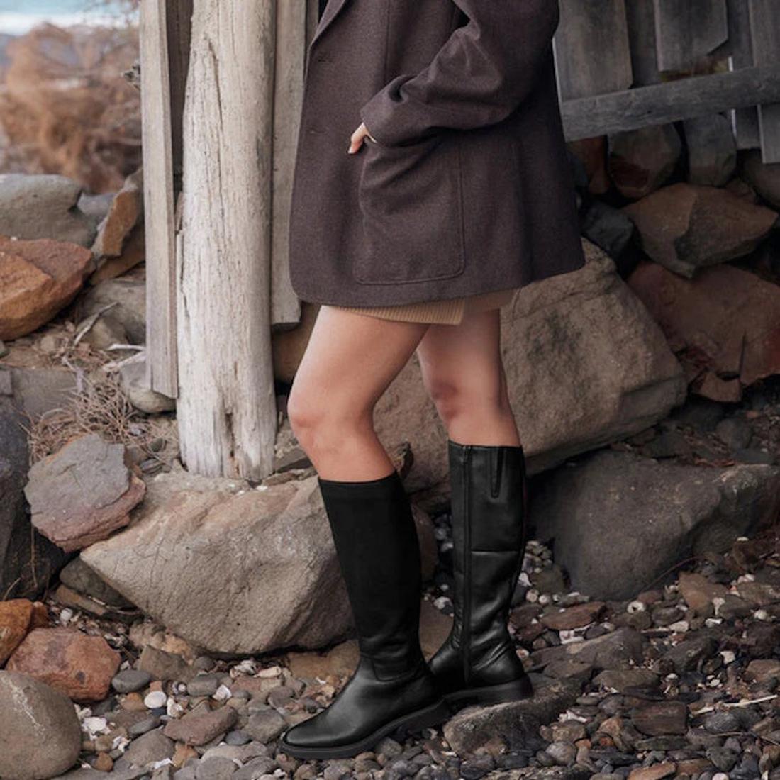 A person stands on rocky ground by a wooden structure, wearing a brown coat, tan skirt, and the EOS Wyoming Black High Leather Womens Heeled Boot. Only their lower body is visible., 9321191201733, 9321191201740, 9321191201757, 9321191201764, 9321191201771