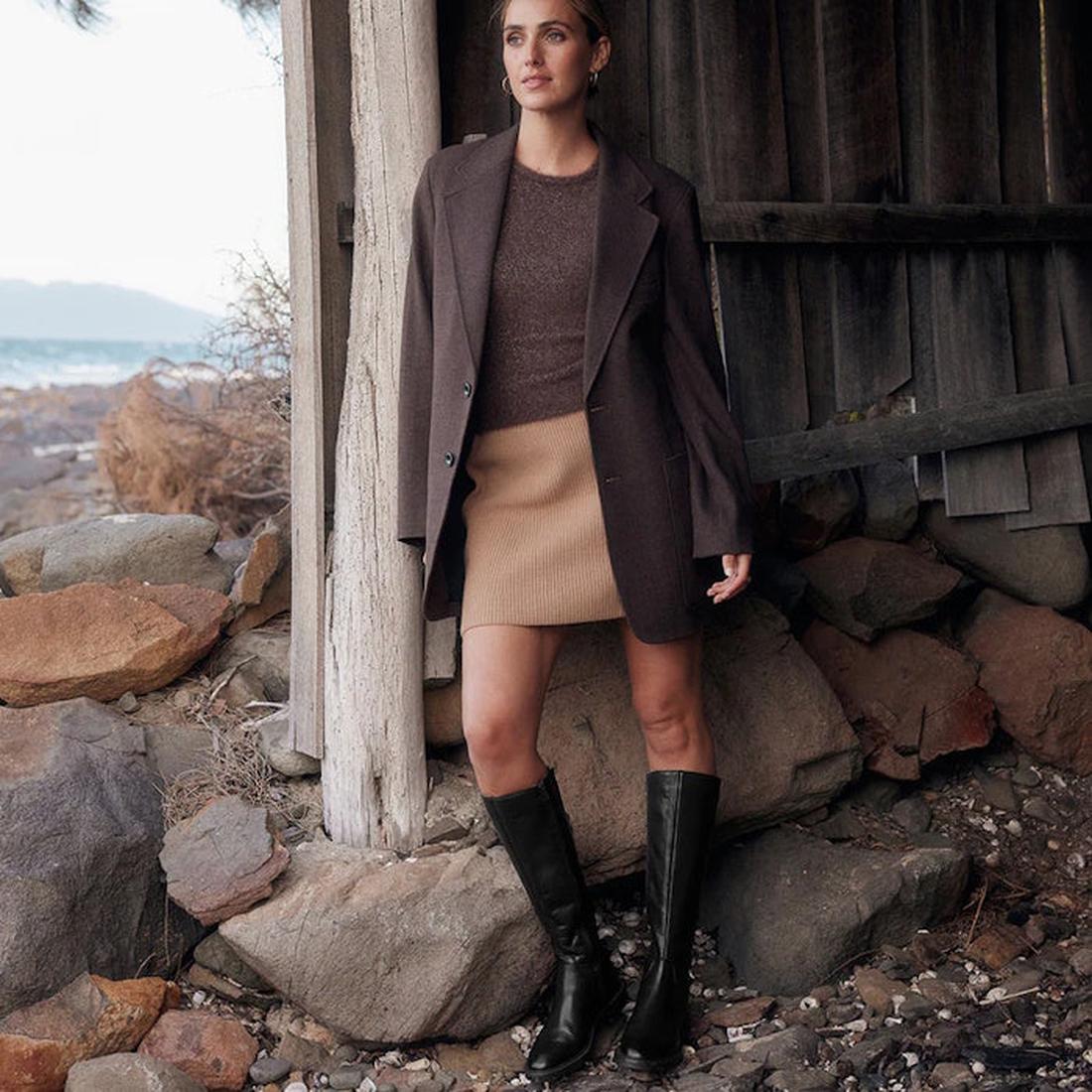A woman stands by a weathered shed on a rocky shore, thoughtfully gazing into the distance. She wears a brown blazer, tan skirt, and completes her look with EOS Blanche Womens Black Leather High Boot Low Heel., 9321191187808, 9321191187815, 9321191187822,