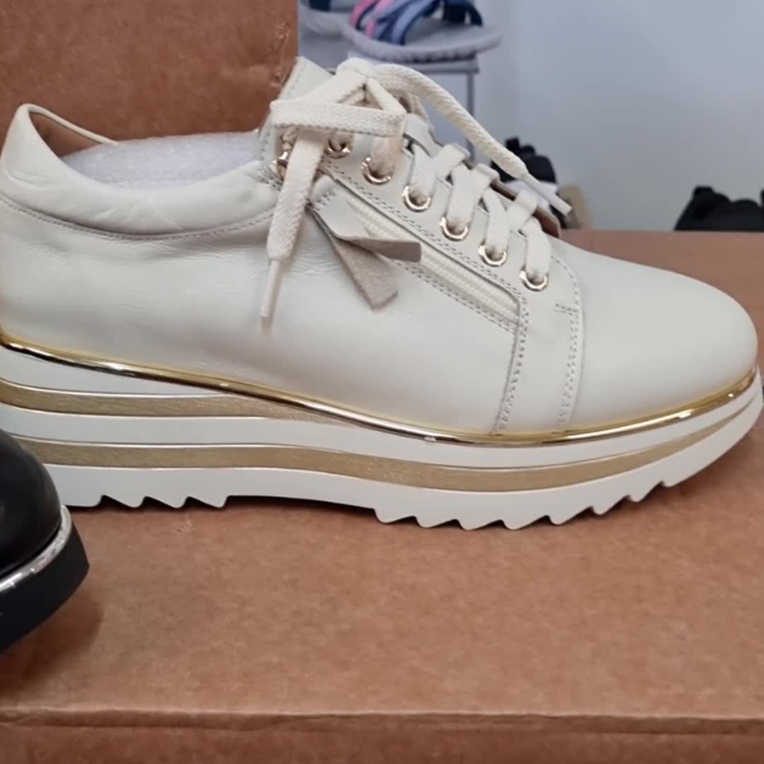 The Alfie & Evie Hosting Womens Leather Shoe in Cream/Gold, featuring a thick platform sole and gold accents, is displayed atop a brown box, with other footwear softly blurred in the background., 38135, 9353738178833, 9353738178840, 9353738178857, 9353738