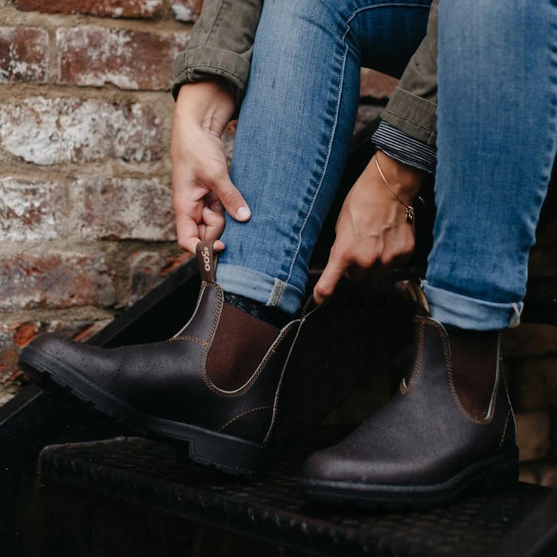 A person sitting down pulling on a right foot of a brown blundtone 500 boot using the pull tabs on the front and the back. Shows the elastic gusset on the inside of the foot, Also shws the left boot on the foot and the person is wearing blue jeans.
931589