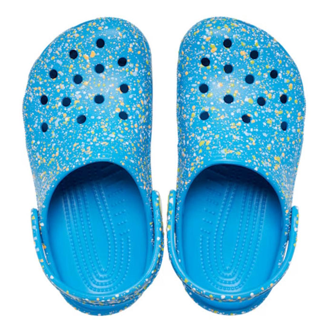 The Crocs Kids - Youths Classic Clog K Paint Splatter Print Blue Multi features blue clogs with yellow and white splatter, top ventilation holes, and adjustable heel straps—a classic kids’ style viewed from above., 198445210136, 198445210143, 198445210150