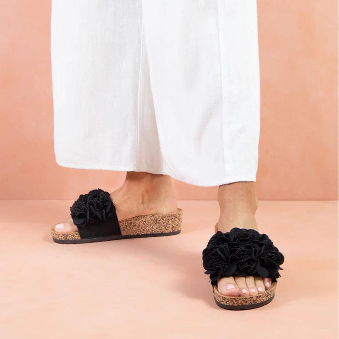 A person wearing white wide-leg pants and Los Cabos Corsage Black Flower Women's Slides, which feature large floral embellishments, stands on a light pink background., 9321191212517, 9321191212524, 9321191212531, 9321191212548, 9321191212555, 46753