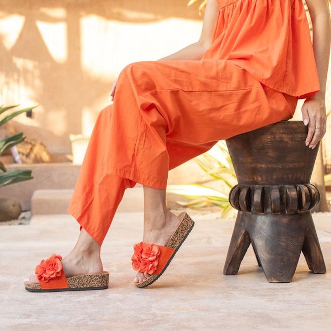 A person in an orange outfit sits on a carved wooden stool, wearing Los Cabos Corsage Tango Peach Flower Women's Slides—featuring orange floral design, cork soles, and a subtle wedge heel—against a sunlit, plant-filled background with earthy tones., 93211