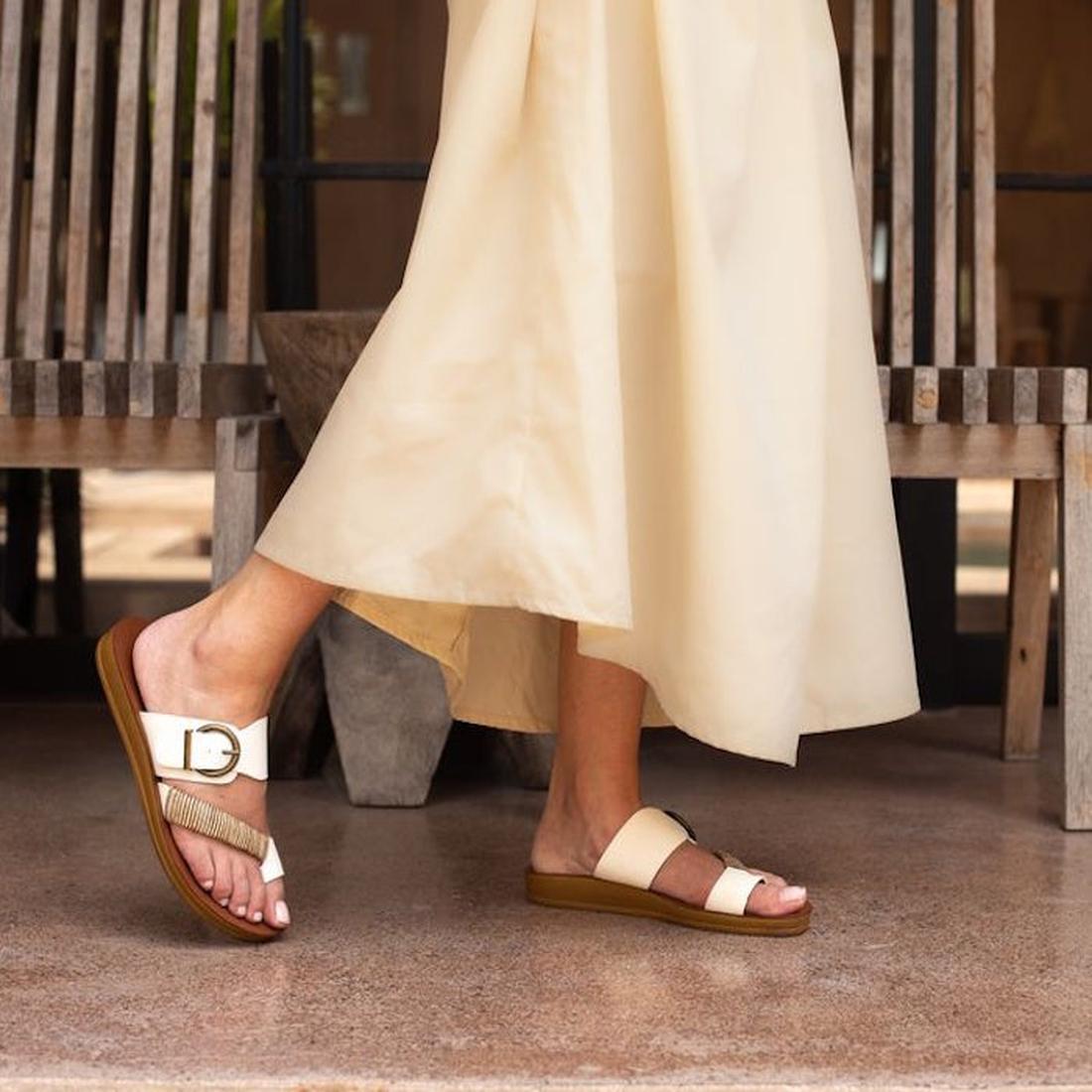 A person wearing a long, cream-colored skirt and Los Cabos Bria Sunset Light Flexible Sandal Jandal walks on a polished floor near wooden chairs, with only the lower part of their body visible., 46928, 9321191296098, 9321191296104, 9321191296111, 93211912