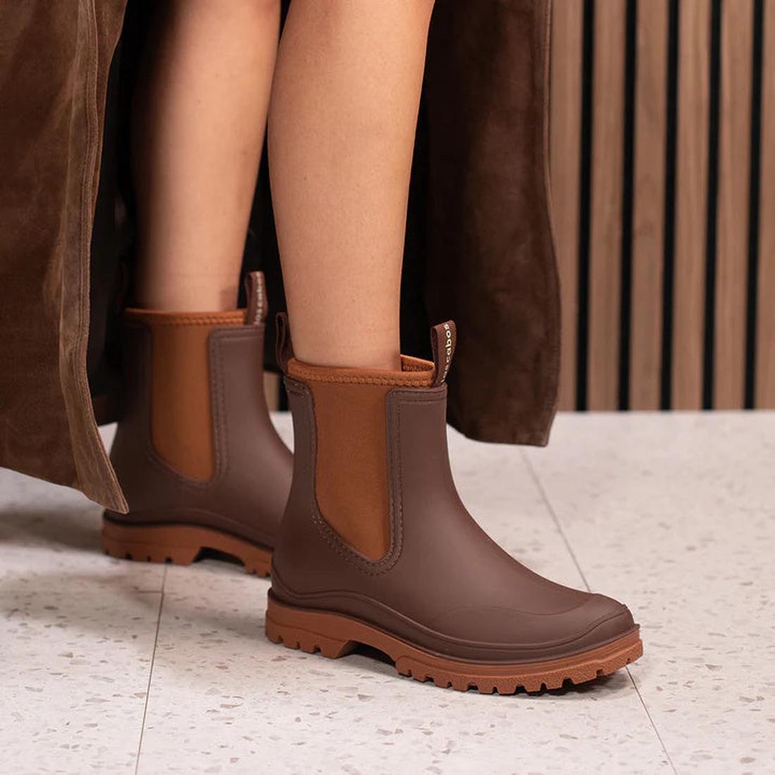 Close-up of a person wearing Los Cabos Amelia Chocolate Ankle Rain Boot Waterproof, featuring rugged soles and a neoprene lining, on a light speckled floor. Part of a brown coat and wooden wall appear in the background., 50781, 9321190004915, 932119000492