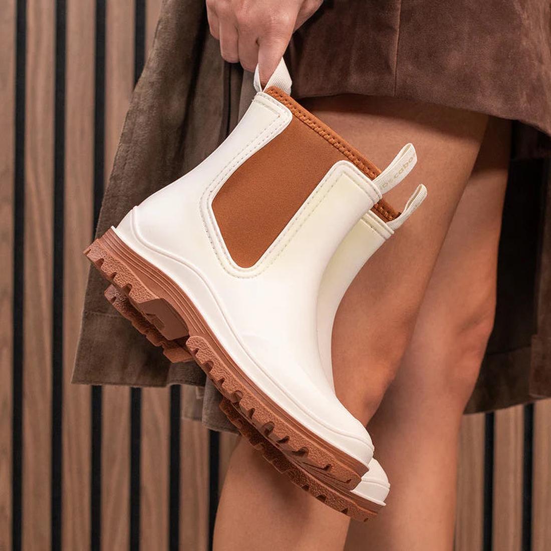 A friendly hand displays the Los Cabos Amelia Cream Ankle Rain Boot—waterproof white with a brown side panel, easy pull-on, sturdy sole.  ksnz, 9321190004977, 9321190004984, 9321190004991, 9321190005004, 9321190005011, 9321190005028, 50794