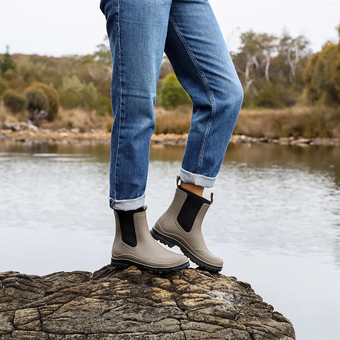 Child wearing Los Cabos Amelia Khaki Ankle Rain Boot—waterproof, slip-on style with sturdy sole—stands on a rock by calm water.  ksnz, 9321191166711, 9321191166728, 9321191166735, 9321191166742, 9321191166759, 9321191166766