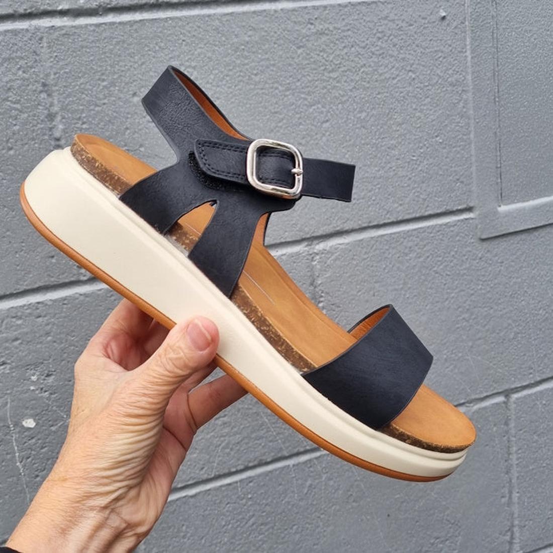 A hand holds the Los Cabos Cartis Black Women's Summer Sandal with a thick white sole, molded footbed, open toe, and silver buckle against a gray textured wall., 9321191298894, 9321191298900, 9321191298917, 9321191298924, 9321191298931, 47459