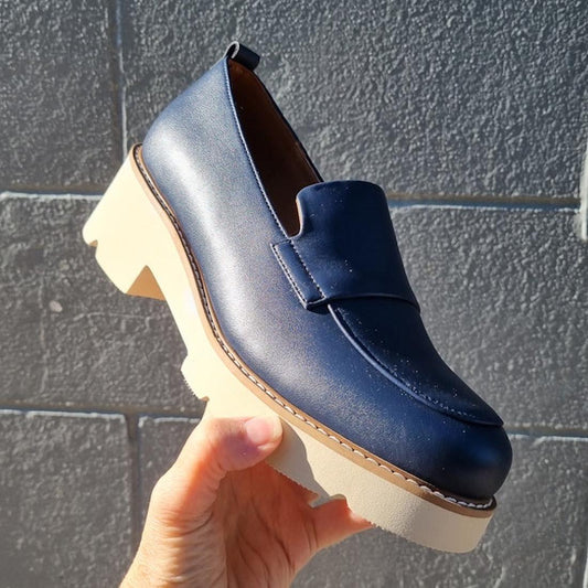 A parent's hand holds the One Trick Pony Newness Navy Leather Women's Chunky Loafer, showing its sturdy sole and easy slip-on design.  ksnz