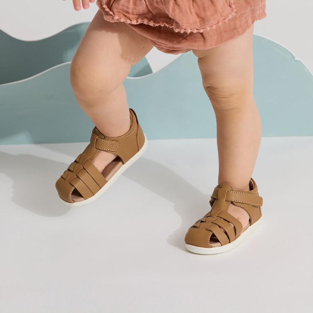 A toddler in a pink outfit wears Pretty Braver Billie First Walker Sandal Closed Back - Tan, standing on a white surface with light blue decorative shapes in the background., 9420075334359, 9420075334342, 9421036357264